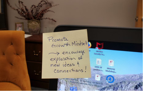 Image showing an office with a sticky note stuck to the corner of a computer screen. The note says "Promote Growth Mindset -- encourage exploration of new ideas & connections"