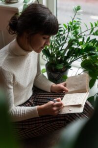 photo of a young woman sitting by a window and writing in a journal