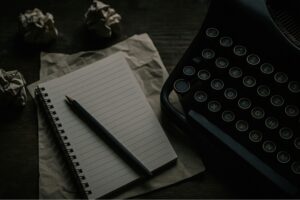 image shows a lined paper with a pencil on it next to a typewriter on a table